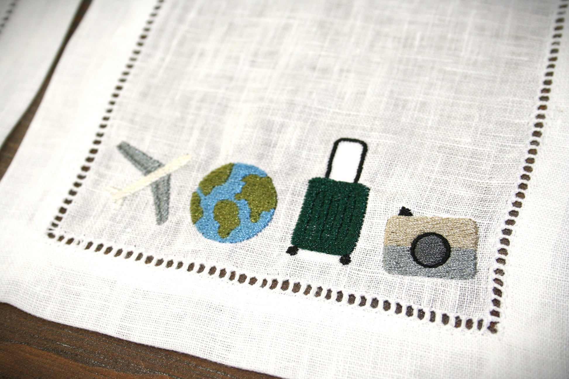 Embroidered linen cocktail napkin featuring an airplane, globe, suitcase and camera for passionate travelers. 