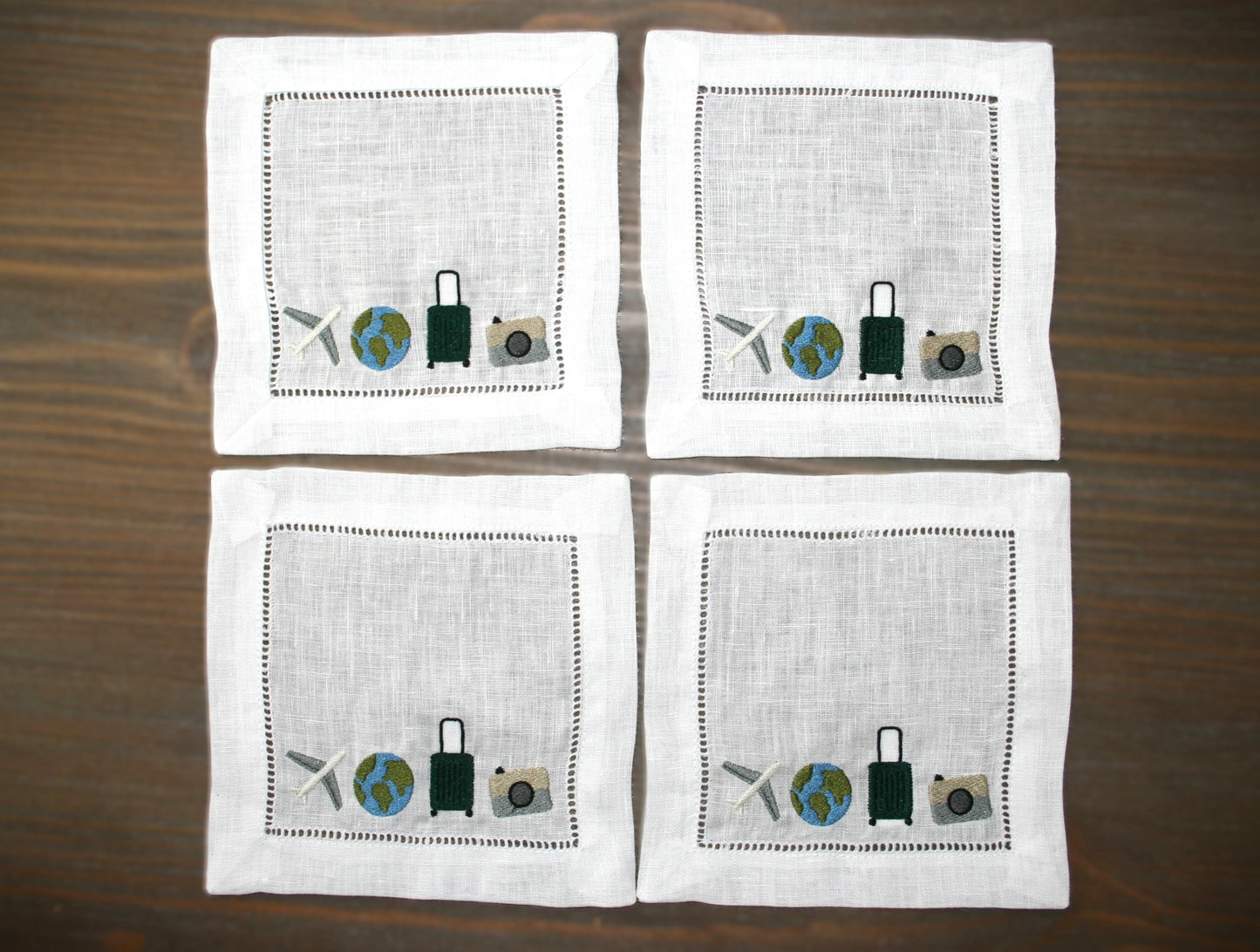 Embroidered linen cocktail napkin featuring an airplane, globe, suitcase and camera for passionate travelers. 