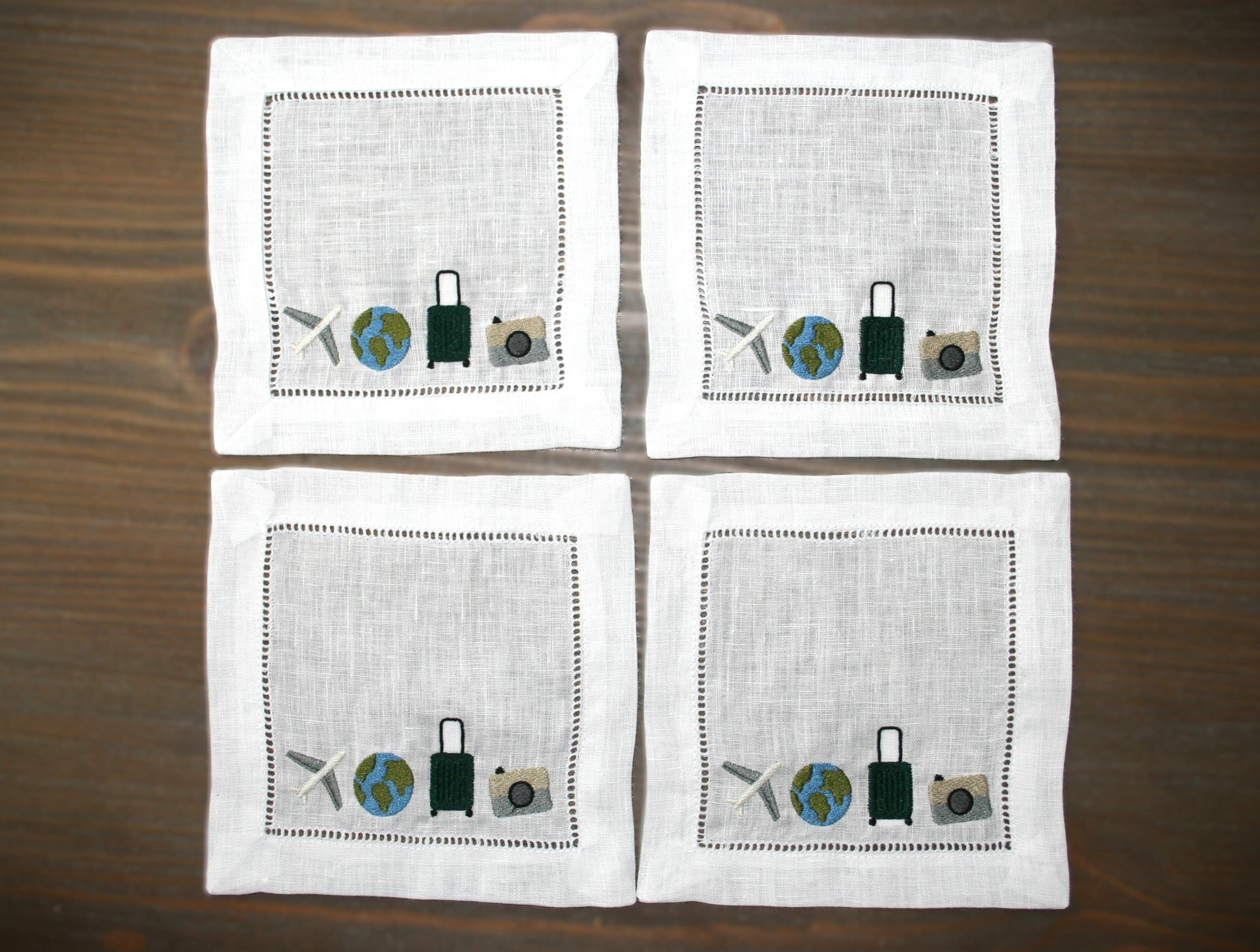 Embroidered linen cocktail napkin featuring an airplane, globe, suitcase and camera for passionate travelers. 